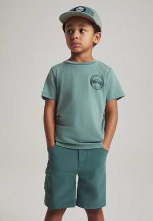 Jeune garçon portant une casquette verte sur le thème de l'aventure, un t-shirt vert assorti et un short, debout les mains dans les poches, regardant sur le côté.
