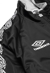 Chaqueta atlética negra con logo blanco. Cuenta con un tejido texturizado y un patrón geométrico en blanco y negro en las mangas. Cierre frontal con cremallera.