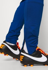 Modré sportovní legíny se zipy na kotnících, které jsou kombinovány s černými fotbalovými kopačkami s oranžovými akcenty a bílým logem ve tvaru swoosh.