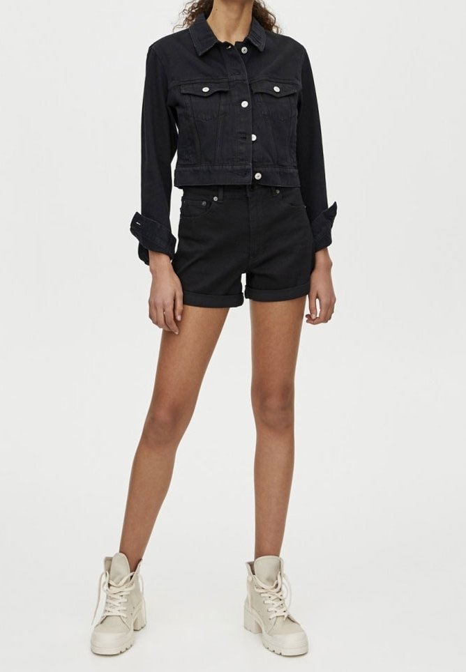 Femme portant une veste en jean noire courte, un short noir à ourlet retroussé et des bottines blanches à lacets, debout devant un fond uni.