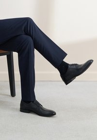 Czarne skórzane buty wizytowe o smukłym kształcie, z przesuwnym zapięciem i niskim obcasem, zestawione z ciemnymi spodniami i wzorzystymi czarnymi skarpetami.