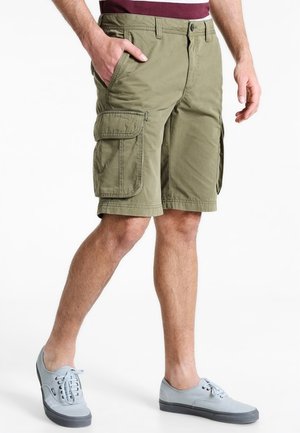 Man draagt olijfgroene cargoshorts en lichtgrijze casual sneakers, staand met één hand in de zak.