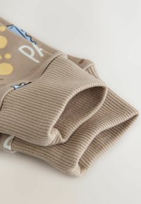 Beige ribstrikkede manchetter på ærmer med delvis tekst og farverige mønstre på lysebrunt stof.