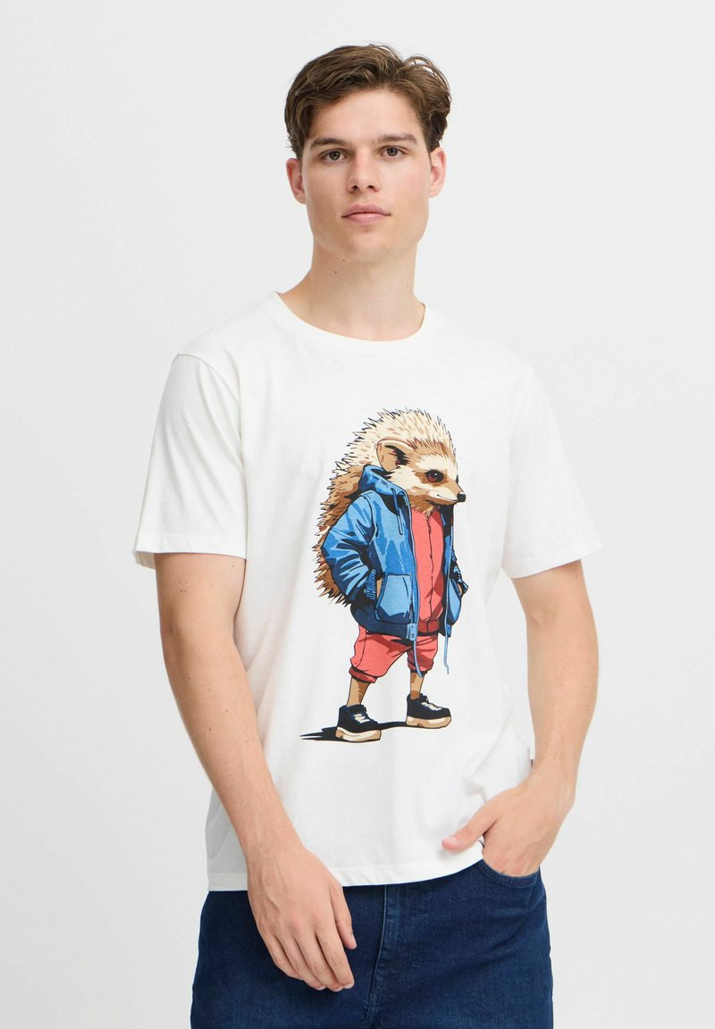 Hvid t-shirt med et tegneserieillustration af et pindsvin iført en blå hættetrøje og røde shorts. Designet inkluderer detaljeret pels og sorte sneakers.