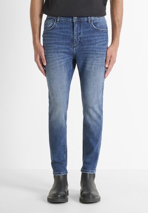 Mężczyzna w wyblakłych niebieskich dopasowanych jeansach i czarnych skórzanych botkach stojący na jednolitym jasnym tle.