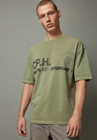 Oliven grøn bomulds t-shirt med korte ærmer og rund halsudskæring. Forsiden har sort tekst og et falmet print. Simpelt, afslappet design.