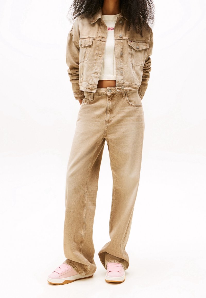 Femme portant une veste en jean beige et un pantalon assorti, avec un top court blanc et des baskets roses, debout les mains dans les poches.