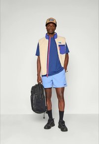 Patagonia fleece vest in crèmekleur met een blauwe zak, gedragen over een blauw shirt en lichtblauwe shorts. Een zwarte rugzak en sneakers maken de look compleet.