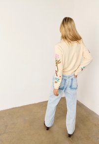 Γυναίκα με μακριά ξανθά μαλλιά στέκεται μπροστά σε έναν λευκό τοίχο, φορώντας ένα εκρού πουλόβερ με πολύχρωμες κεντητές μανσέτες και ανοιχτό μπλε τζιν.