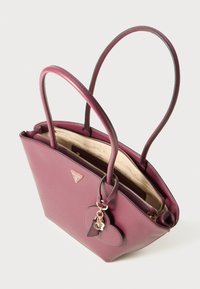 Lilla lærveske med doble håndtak, hjerteformet anheng, glitrende gullfarget glidelås og beige innvendig foring merket med "Guess."