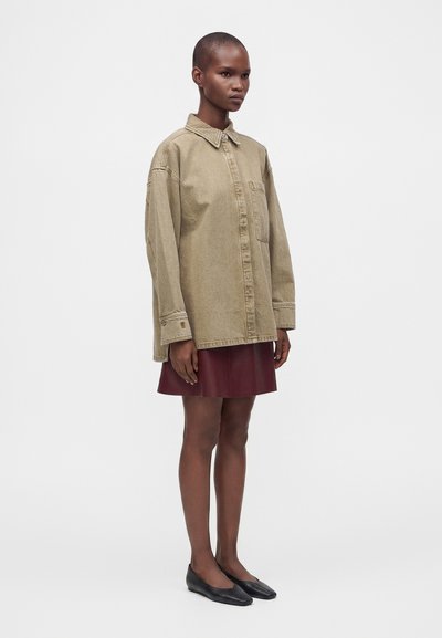 Μοντέλο στέκεται φορώντας μία oversized μπεζ πουκάμισο με κουμπιά, μπορντό δερμάτινη φούστα και μαύρα επίπεδα παπούτσια σε απλό ανοιχτό φόντο.