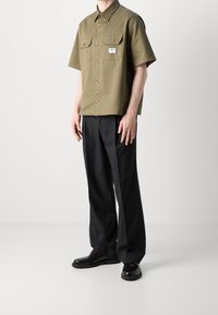 Olivgrünes Kurzarmhemd mit zwei Brusttaschen, kombiniert mit schwarzen maßgeschneiderten Hosen und schwarzen Schuhen mit einer strukturierten Sohle.