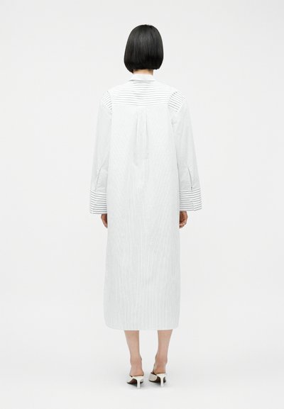Γυναίκα με κοντά μαύρα μαλλιά που φοράει λευκό μεσαίο πουκάμισο φόρεμα με κάθετες και οριζόντιες λεπτές ρίγες, στέκεται με την πλάτη στραμμένη προς τα εμπρός.
