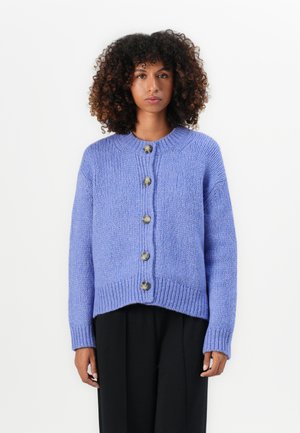 Cardigan lavorato a maglia di un leggero colore viola con scollo rotondo, cinque grandi bottoni e polsini e orlo a coste, abbinato a pantaloni neri.