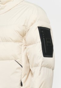 Manche de veste matelassée beige clair avec une poche zippée noire et un écusson portant le logo "BOSS".
