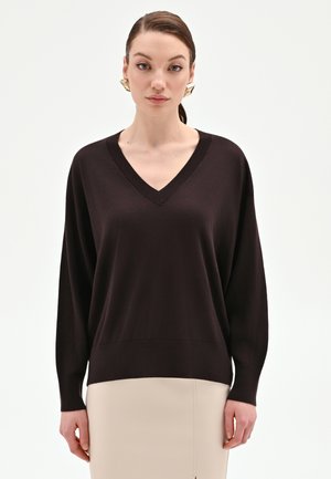 Donna che indossa un maglione scollo a V marrone scuro e una gonna beige, con grandi orecchini geometrici dorati, in piedi davanti a uno sfondo bianco semplice.