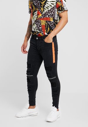 Mężczyzna w czarnych, podartych obcisłych dżinsach z pomarańczowym paskiem po boku, kolorowej, wzorzystej koszuli z krótkim rękawem i białych tenisówkach, stojący z jedną ręką w kieszeni.