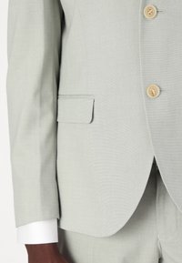 Šviesiai pilkas švarkas su pasirinktiniu atlapu, dviem priekiniais mygtukais ir krūtinės kišene. Yra balti rankogaliai ir tekstūruota medžiaga.