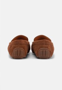 Καφέ σουέντ μοκασίνια, εμφανιζόμενα από πίσω, που αναδεικνύουν την υφασμένη λαστιχένια φτέρνα και τις ραμμένες λεπτομέρειες πάνω σε απλό ανοιχτό φόντο.