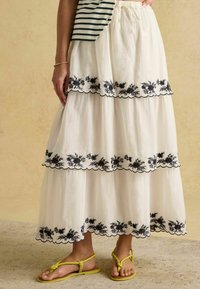 Biela viacvrstvová sukňa s čiernou kvetinovou výšivkou a zaoblenými okrajmi, kombinovaná so žltými sandálmi na remienky. Bavlnený materiál, neformálny dizajn.
