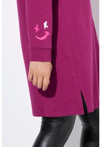 Tunique fuchsia au coupe décontractée, mettant en avant des graphismes de smileys blancs et roses sur les manches. Détail d fente sur les côtés pour plus de liberté de mouvement.