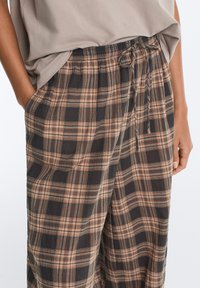 Personne portant un pantalon ample à carreaux avec des poches latérales et une chemise beige légèrement rentrée à la taille.