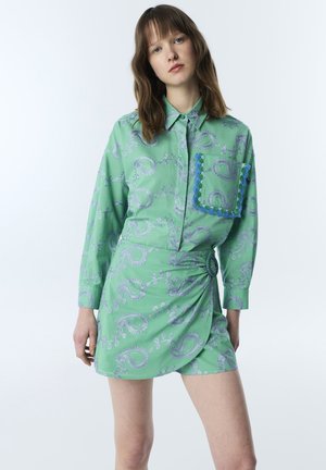 Vrouw draagt een groen paisley patroon blouse met lange mouwen, een grote zak en bijpassende high-waist minirok met zijsluiting.