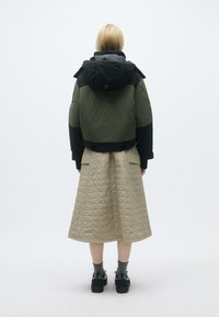 Jaqueta verde e preta curta com um grande capuz, combinada com uma saia acolchoada bege. O calçado é preto, com meias cinzas. Design simples e funcional.