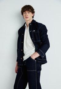 Young man with curly hair wearing dark denim jacket and pants with visible white stitching, white shirt, standing against plain wall.