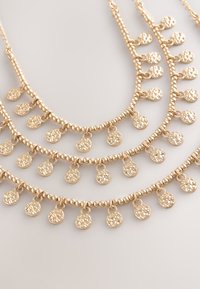 Vier goldene Halsketten mit kleinen, runden, gehämmerten Anhängern, die gleichmäßig entlang von Perlenketten auf hellem grauem Hintergrund angeordnet sind.