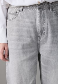 Jeans de denim gris claro con una textura suave, que cuentan con un diseño de cinco bolsillos, presillas para cinturón y una cremallera con cierre de botón.