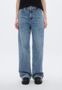 Jean large bleu clair en denim. Caractérisé par une taille haute, des poches avant et une fermeture classique à bouton. Associé à des chaussures noires.