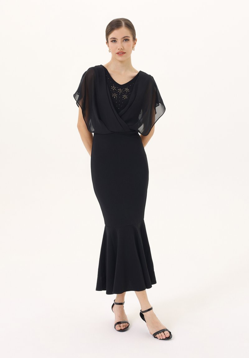 Czarna wieczorowa sukienka o dopasowanej sylwetce i rozszerzanym dołem. Przezroczysta nakładka na ramionach, zdobiony dekolt koralikami. Połączona z czarnymi sandałami.