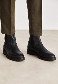 Bottines chelsea en cuir noir avec semelles en caoutchouc, dotées de panneaux latéraux élastiques et d'un bout arrondi, portées avec un pantalon vert olive.