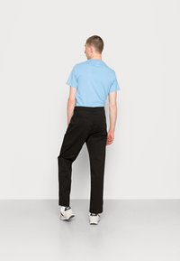 Lässiges Männeroutfit mit einem hellblauen Kurzarmhemd und schwarzen Hosen, mit Gesäßtaschen und weißen Sneakers. Neutraler Hintergrund.