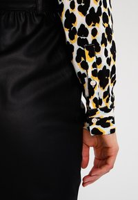Camisa de tela con estampado de leopardo y patrones coloridos, con un puño de botón negro. Combinada con una falda de cuero negro lisa.