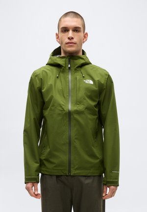 Férfi olívazöld vízálló kabátot visel kapucnival, teljes elülső cipzárral és logóval a mellkason, sima háttér előtt áll.