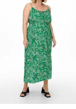 Vrouw draagt een groene mouwloze maxi-jurk met een paisley-patroon en zwarte sandalen met hak.