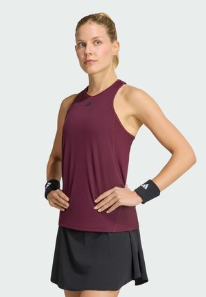 Donna atletica che indossa un top bordeaux senza maniche, gonna nera e polsini neri con loghi, in piedi con le mani sui fianchi davanti a uno sfondo semplice.
