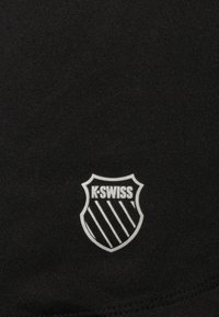 Tissu noir avec une surface texturée arborant un logo de bouclier K-Swiss blanc, accentué par des bandes diagonales et une typographie en gras.