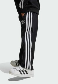 Pantalons de sport noirs avec trois bandes blanches sur les côtés et un logo Adidas sur la hanche, assortis à des baskets blanches avec des accents noirs.