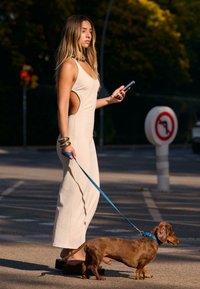 Žena u bež haljini s izrezima i debelim trakama, drži pametni telefon dok šeta smešnim dobermanom na plavoj uzici, noseći platforme.