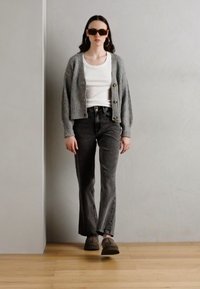 Grå ribbet cardigan med knapper, hvit skjorte, mørkegrå flared jeans, brune sko og overdimensionerte solbriller på en person som står mot en vegg.