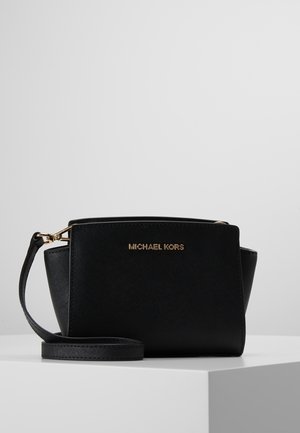 Sort Michael Kors håndledsrem med gyldent logo og aftagelig rem vist på hvid overflade mod grå baggrund.