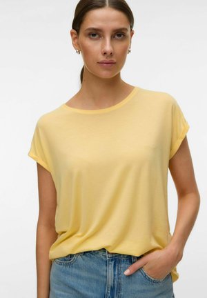 Woman wearing a light yellow short-sleeve top tucked into blue denim jeans, with one hand in pocket, facing front on plain background.