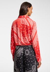 Blusa rossa con motivo a fantasia, a maniche lunghe, caratterizzata da un design paisley e accenti floreali. Il tessuto sembra morbido e leggero.