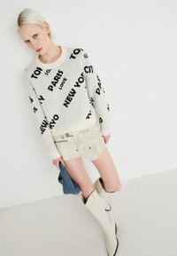 Kremgenser med svarte bynavn, beige semsket skinn mini nederdel, og beige støvler til kneet. Glatte teksturer, tettsittende design, og kontrasterende accenter.