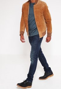 Béžová semišová bomber bunda s žebrovanými manžetami, modrá texturovaná košile, tmavé slim-fit džíny a černé šněrovací boty s hnědou podrážkou.