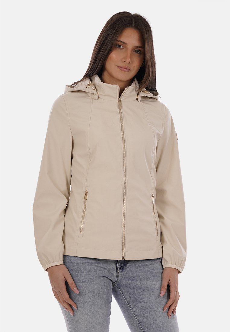 Donna che indossa una giacca beige con cerniera, cappuccio e tasche laterali con cerniera, abbinata a jeans azzurri, in piedi davanti a uno sfondo semplice.