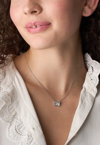 Jonge vrouw met een zilveren vlinderhanger, kleine zilveren oorbel en een witte blouse met een kanten kraag.